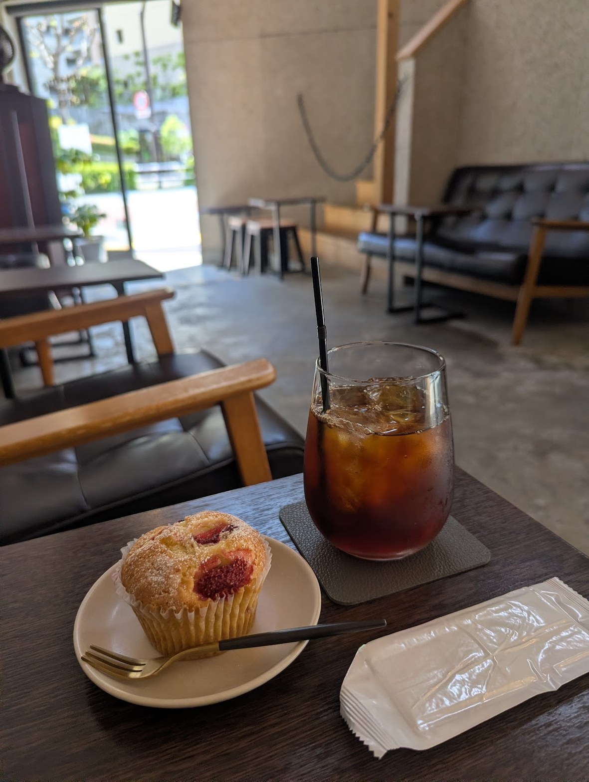 ベリー入りマフィンと氷入りアイスコーヒーがテーブルに置かれ、奥にソファ席と窓が見える店内。